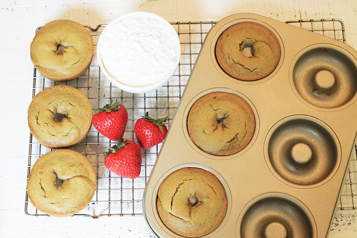 Donuts with bowl of powdered sugar and strawberries on a cooling rack