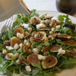 Fresh Fig and Goat Cheese Over Arugula with chopped pecans, balsamic dressing being poured on it.