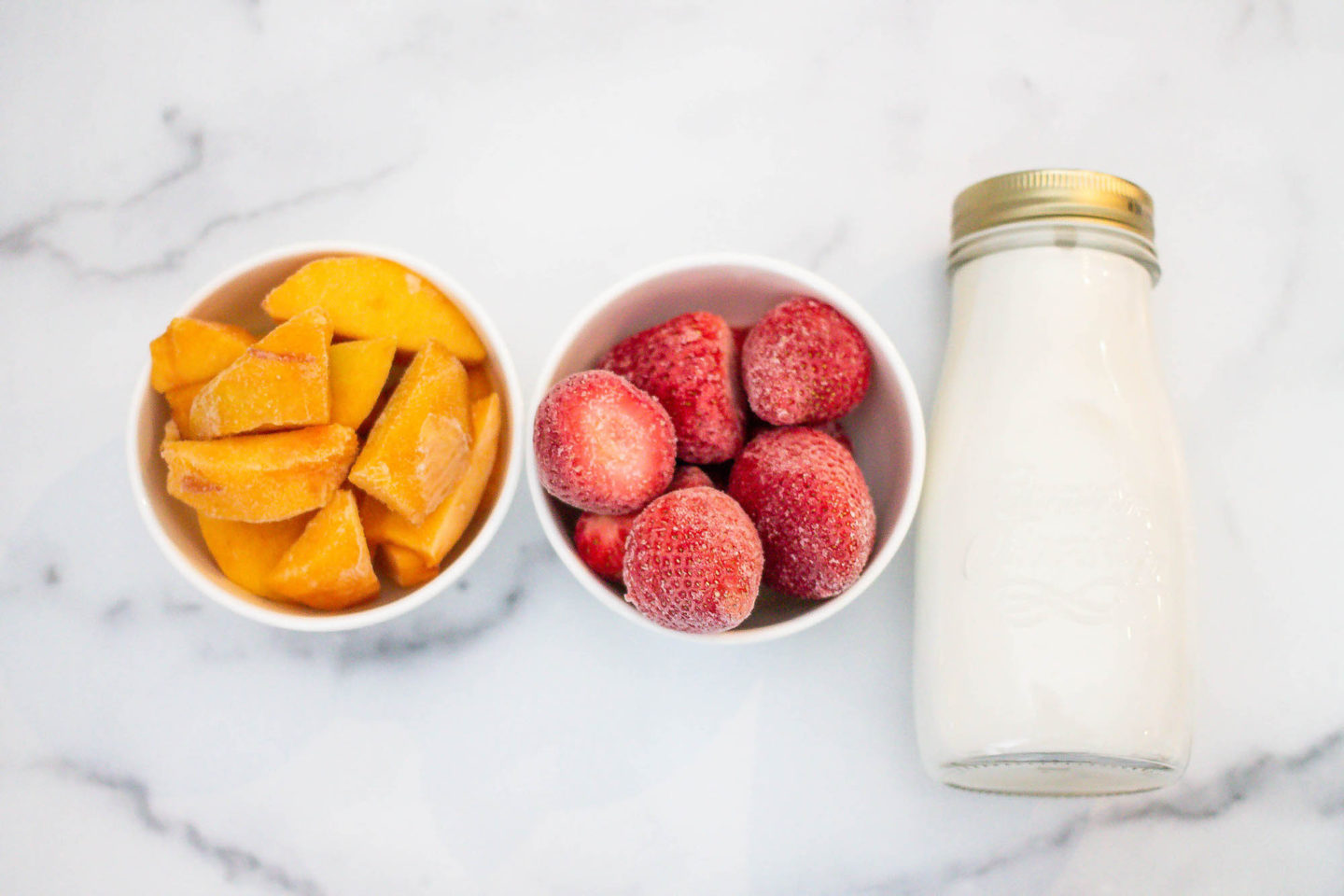 Frozen chopped peaches in a bowl and frozen strawberries in a bowl and almond milk in a glass jar, ingredients for the Peach and Strawberry Smoothie Bowl