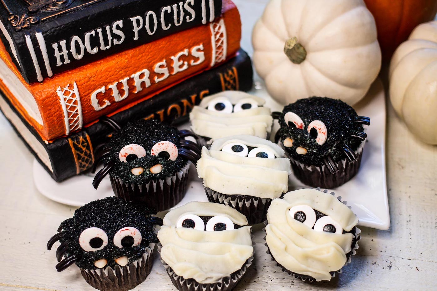 Chocolate cupcakes decorated like spiders and mummies on a white platter with Halloween decorations and small pumpkins