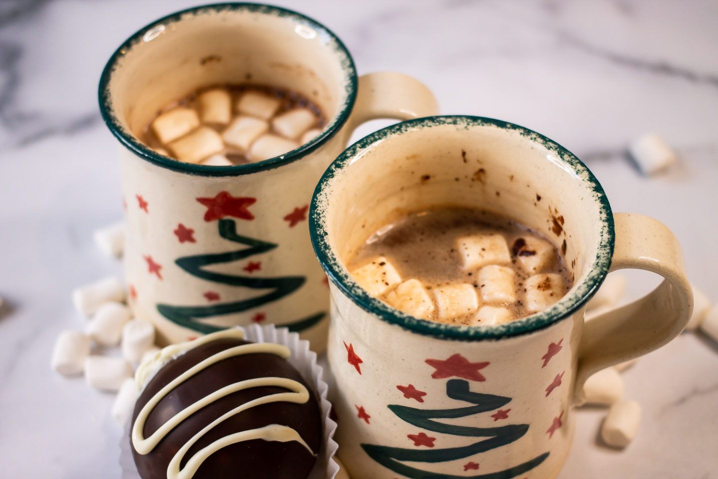 Two cups of hot cocoa with mini marshmallows and one hot cocoa bomb in a cupcake liner
