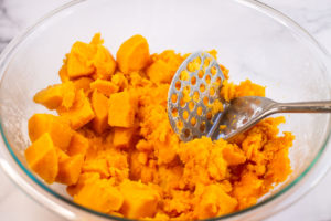 Glass bowl of cooked sweet potatoes, partially mashed with potato masher